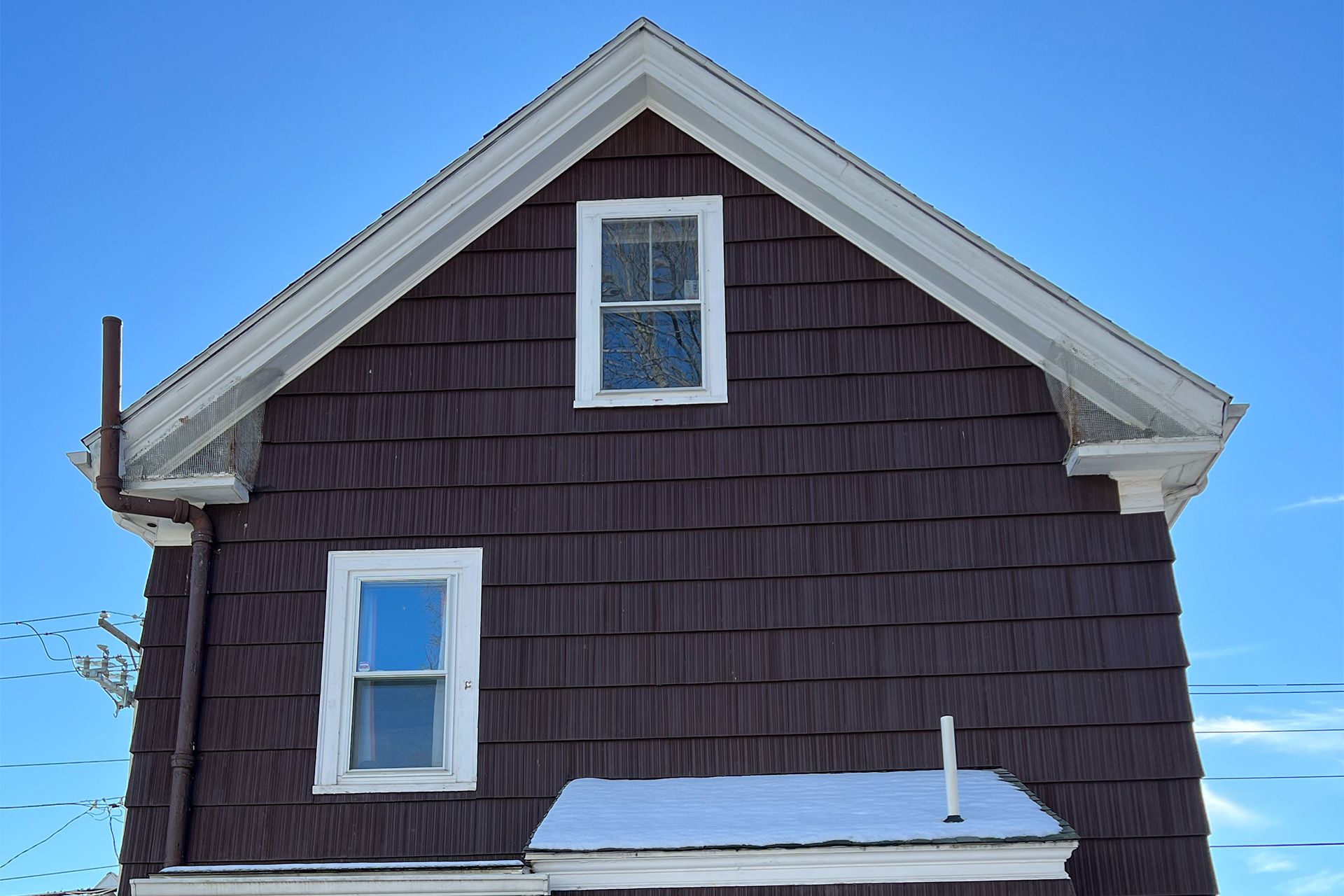 Dark-shingled house side with white-trimmed windows and snow on the roof under a blue sky