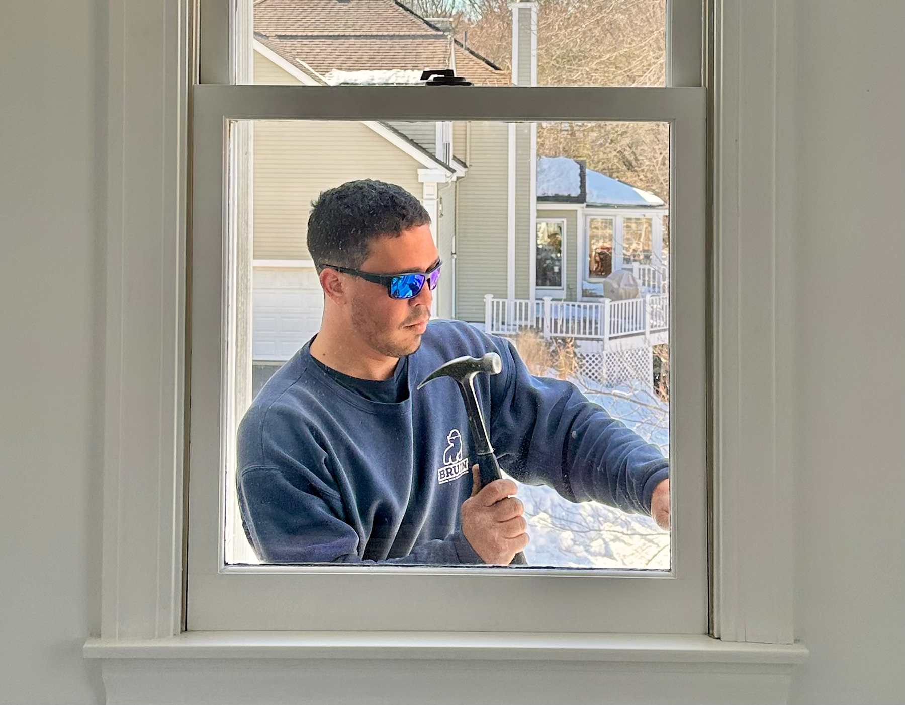 Man in blue jacket working outside a window, holding a hammer and wearing sunglasses.