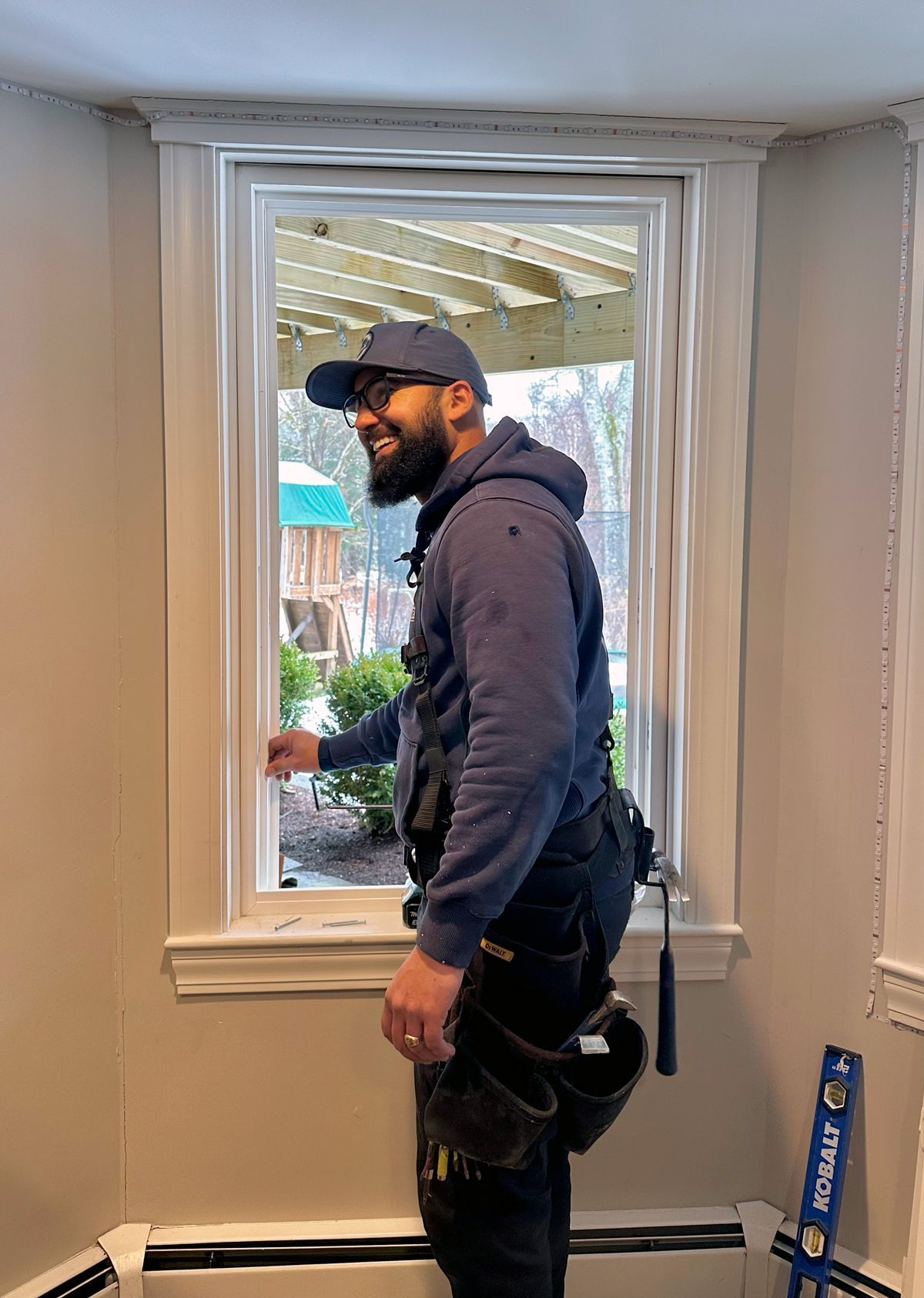 Man standing indoors by a window, wearing a cap and work harness, with tools nearby.