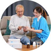 Caregiver in blue handing medication to smiling man seated on a couch, with pill boxes and medicine bottles nearby