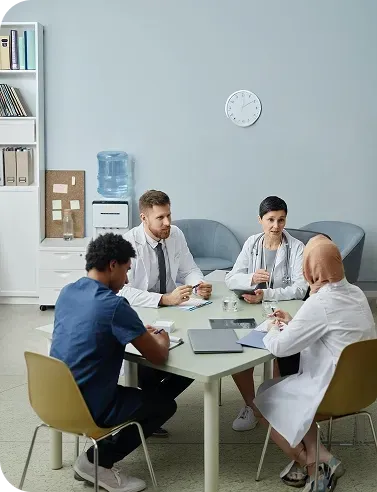 Medical team meeting around a table in a bright office, with laptop, papers, and coffee mugs.