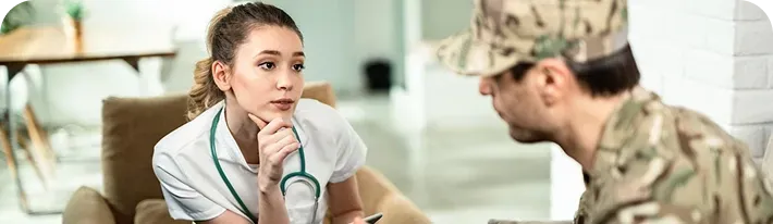 Woman in white top speaking with a soldier in camouflage, seated indoors with a serious expression.