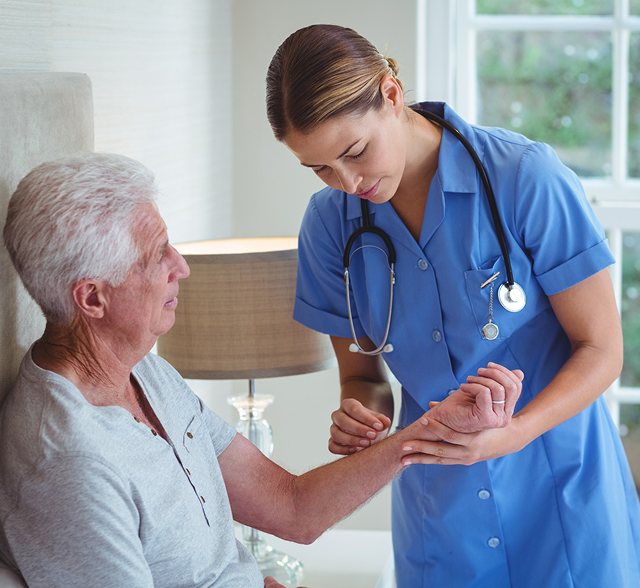 Nurse in blue scrubs checks an elderly patient’s wrist in a bright home setting.