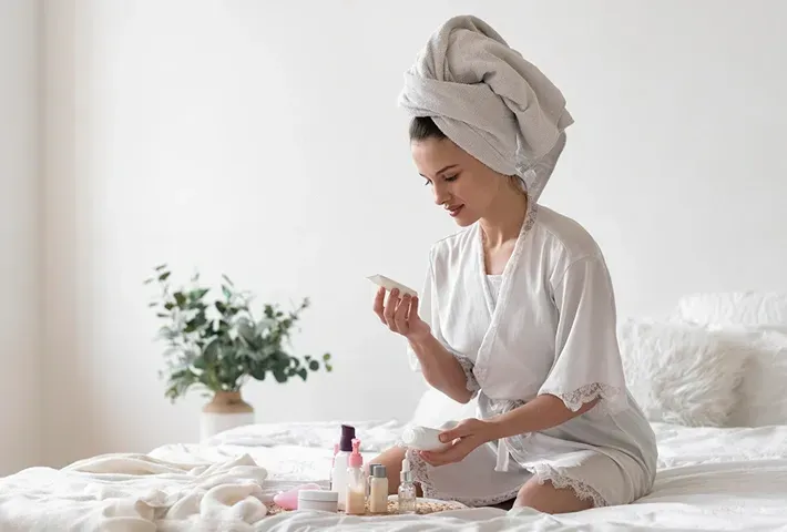 Woman applying skincare on a bed with towel wrapped around her head in a bright, cozy bedroom