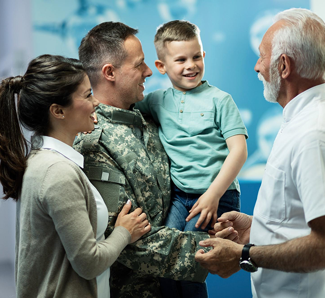 A soldier holds a smiling child while shaking hands with an older man; a woman stands beside them indoors.