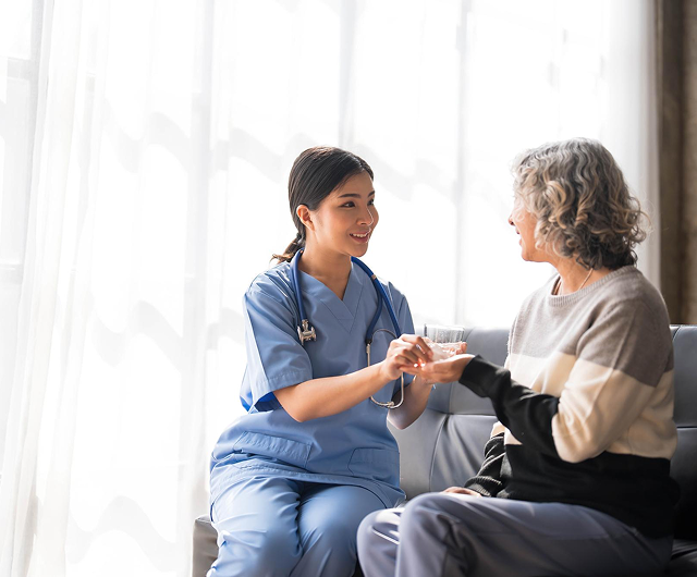 Nurse in blue scrubs talking with an older patient on a couch in a bright room