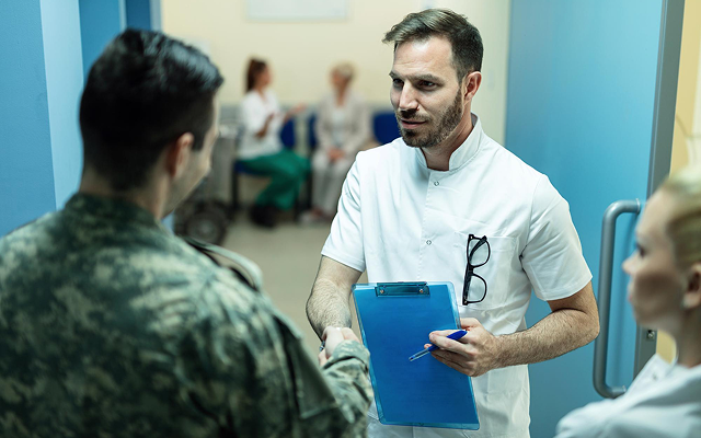 Doctor holding a blue clipboard talks with a military patient in a clinic hallway