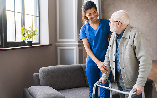 Caregiver in blue scrubs helping an elderly man with a walker in a bright room