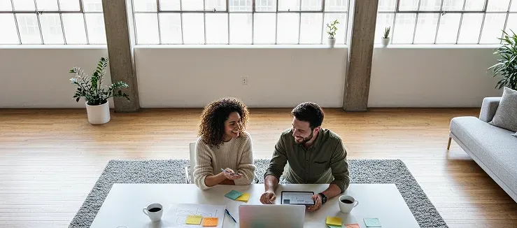 Two people sitting on the floor at a coffee table, working on a laptop in a bright living room