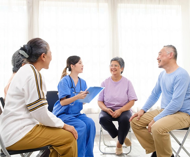 Group therapy session with four people seated in a bright room, talking with a counselor holding notes