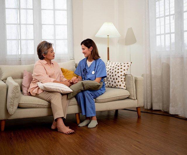 Woman in blue scrubs sitting with an elderly woman on a sofa in a bright living room