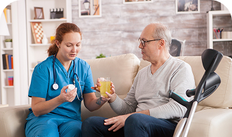 Nurse in blue scrubs talking with a man in a wheelchair, both holding drinks in a bright living room.