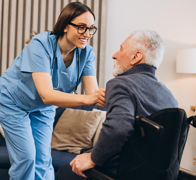 Care worker in blue scrubs helps seated resident in a wheelchair in a bright living room