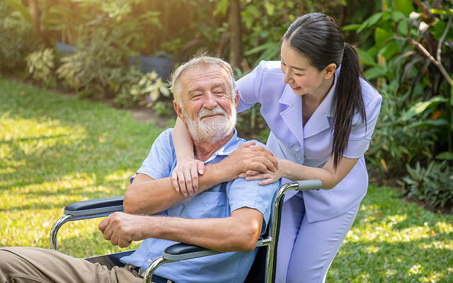 Caregiver in light purple uniform smiling and helping a man in a wheelchair outdoors in a sunny garden