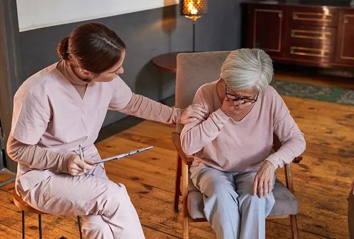 Two women in a living room, one helping the other from a chair with a cane nearby.
