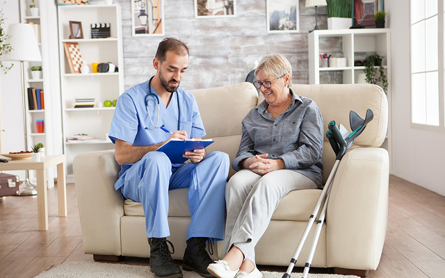 Nurse showing notes to a seated patient on a couch in a bright living room, with a pair of crutches nearby