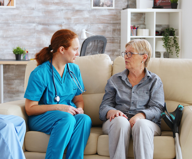 Caregiver in blue scrubs talks with an older person on a beige couch in a bright living room