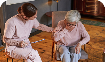Caregiver helps seated woman with exercise bands in a warm living room