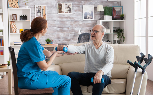 Caregiver in blue scrubs checks blood pressure of seated patient in a living room