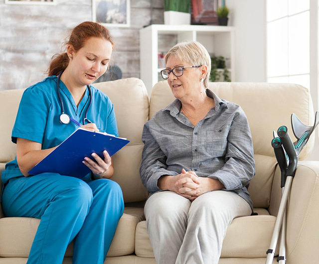 Caregiver in blue scrubs taking notes with an older woman on a couch, with a walker beside them.