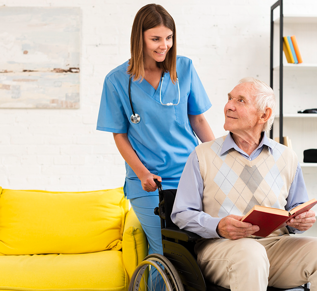Nurse in blue scrubs speaking with a man in a wheelchair beside a yellow sofa.