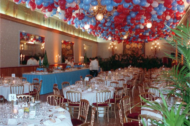 Salle de banquet décorée avec un plafond de ballons rouges et bleus, des nappes blanches et des tables rondes soigneusement dressées.