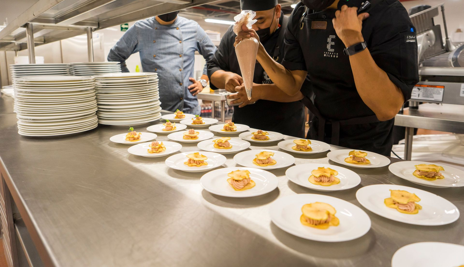 Le personnel du traiteur dresse des assiettes d'amuse-gueules sur des plats blancs, devant un comptoir de cuisine en acier inoxydable.