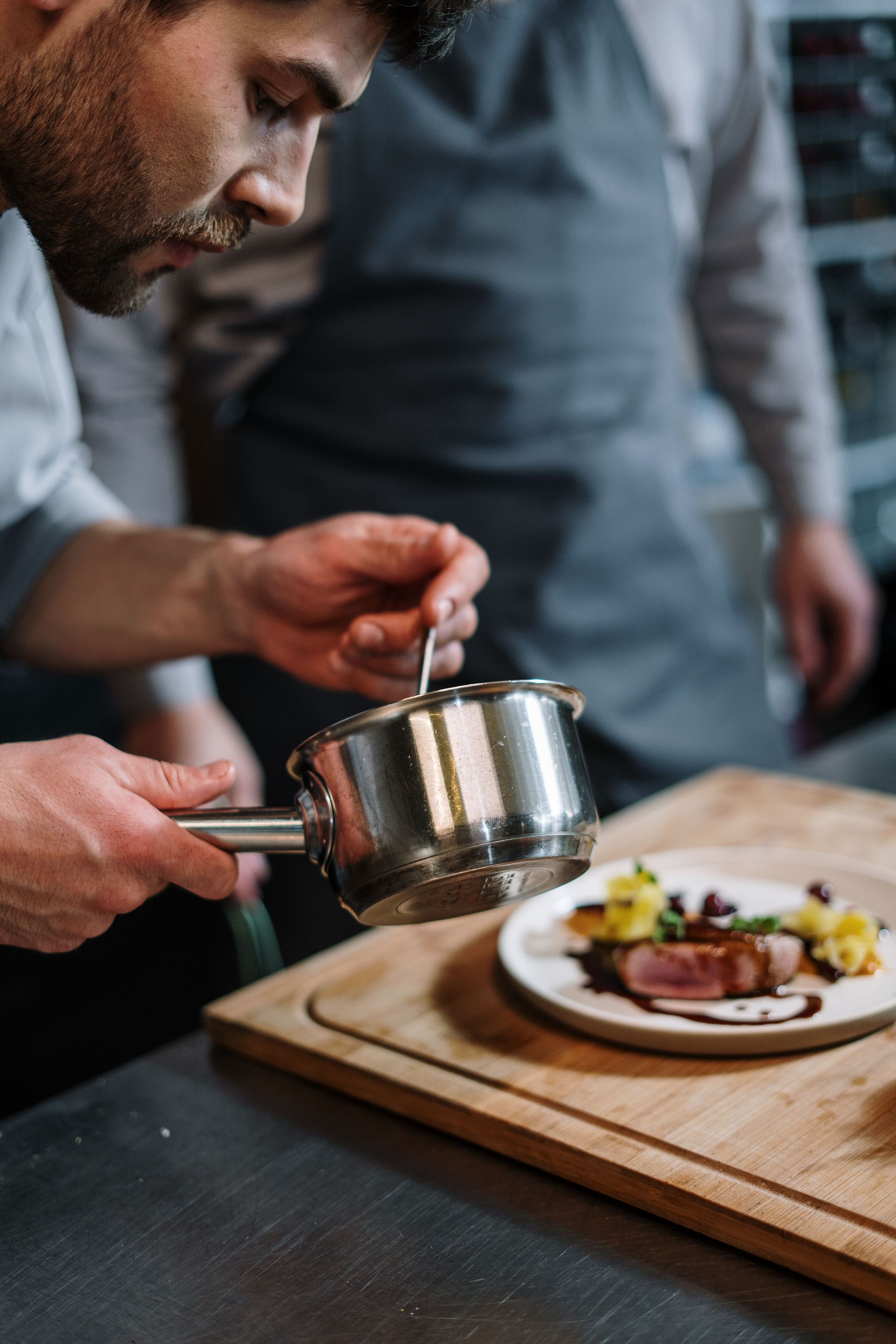 Un chef dresse un plat avec une petite casserole posée sur une planche en bois dans une cuisine.