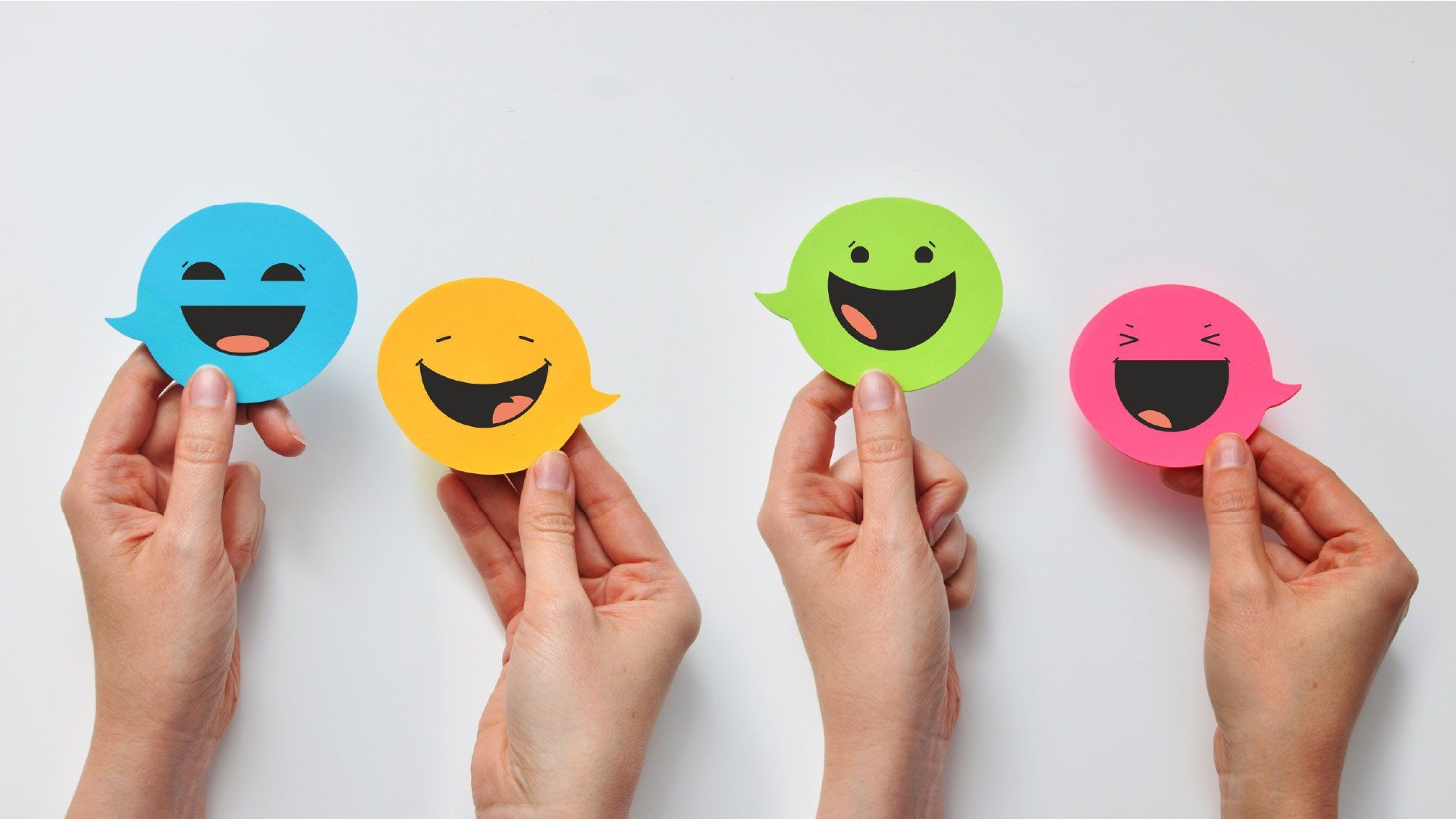 Four hands holding colorful smiling paper face cutouts against a light background