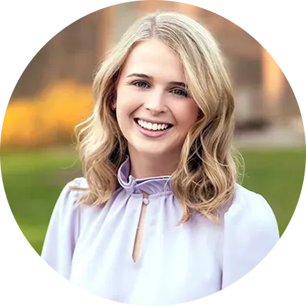 Woman with blonde wavy hair, smiling, wearing a light purple blouse outdoors with a green background.