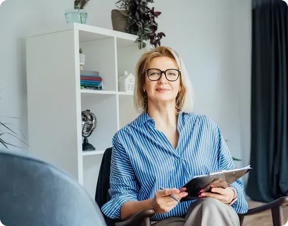 Woman with glasses in a striped shirt, holding a notepad, seated in an office.