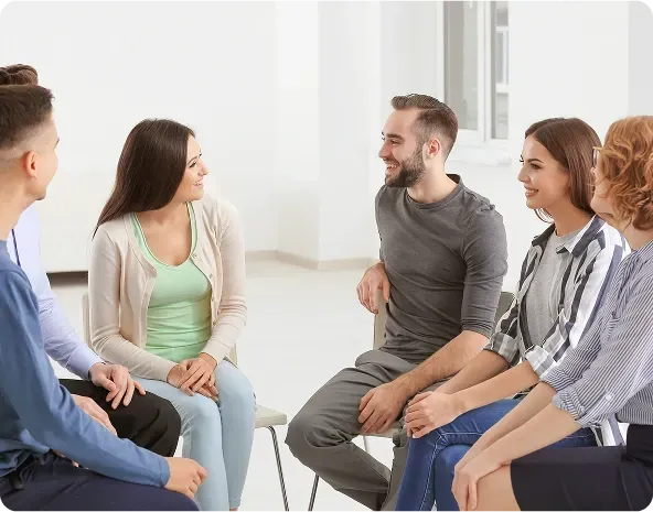 People in a circle, smiling, talking, likely in a support group setting. Light room, chairs, casual clothes.