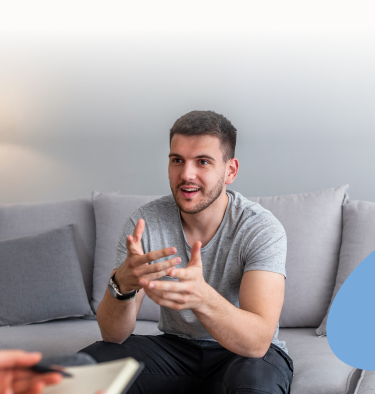 Man gesturing while sitting on a couch, talking to someone off-screen in a counseling setting.