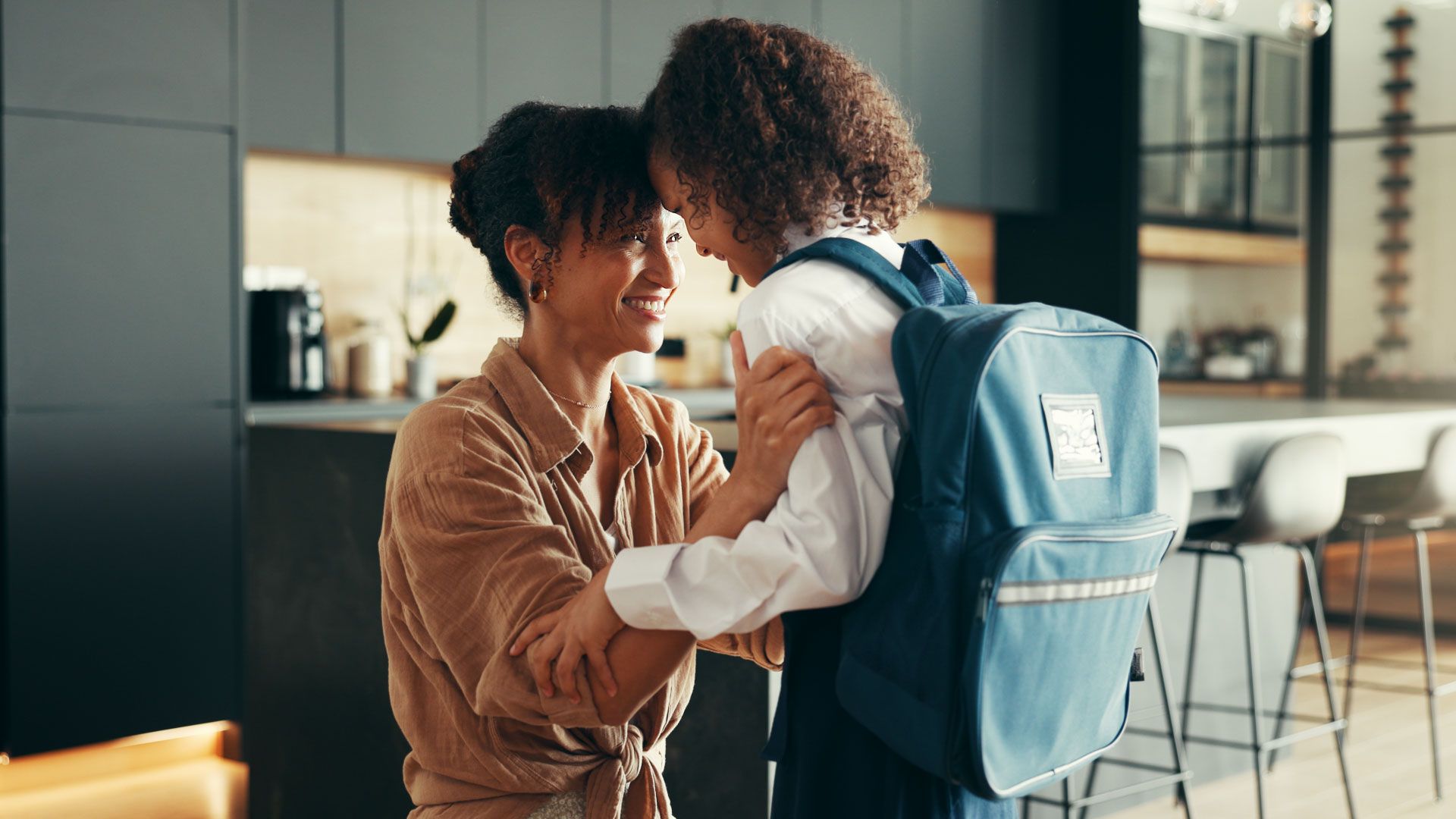 Two people smiling and holding each other in a bright kitchen, one wearing a backpack.