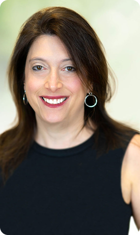 Woman with dark hair smiles, wearing black top, silver earrings, in a softly lit setting.