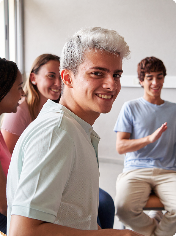 Young man with white-blond hair smiles, looking over shoulder, surrounded by people in light-colored room.