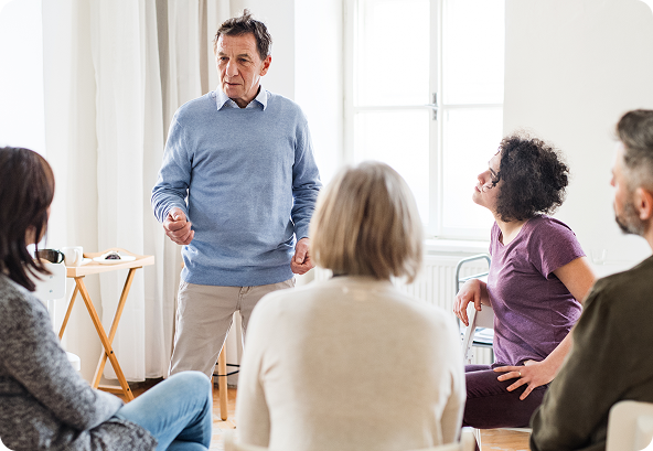 Group therapy session: Man standing, speaking to seated individuals. Light room, chairs, and expressions of listening.