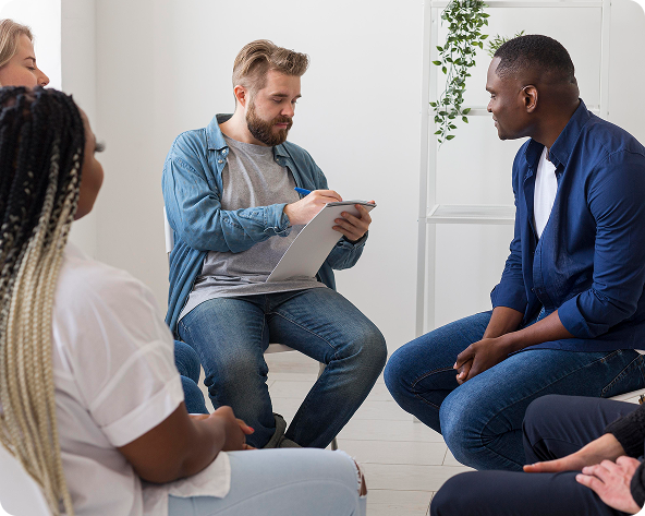 People in a circle during a therapy session; a therapist takes notes while listening to a client.