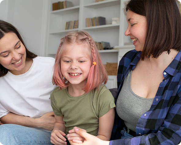Two women and a child laughing on a couch. The child has pink hair and holds hands.