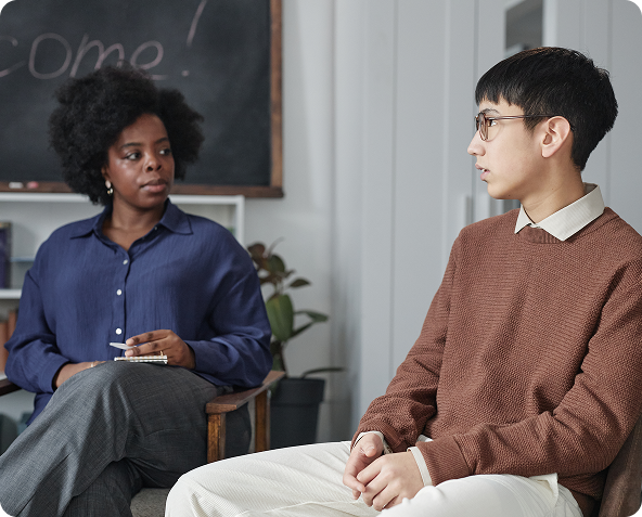 A woman in blue shirt and a person in brown sweater sit facing each other in a room, conversing.