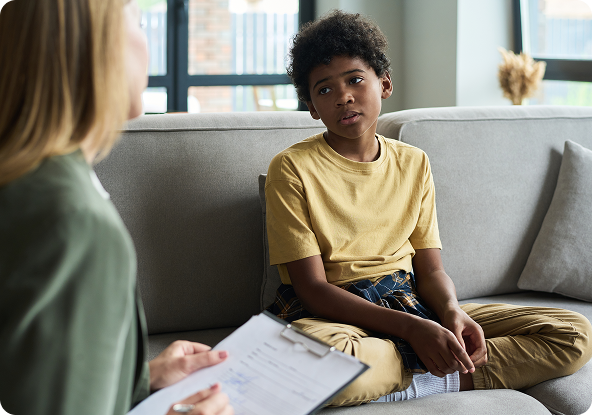 Boy in yellow shirt looks concerned, seated on a couch, facing a person holding a clipboard in a counseling session.