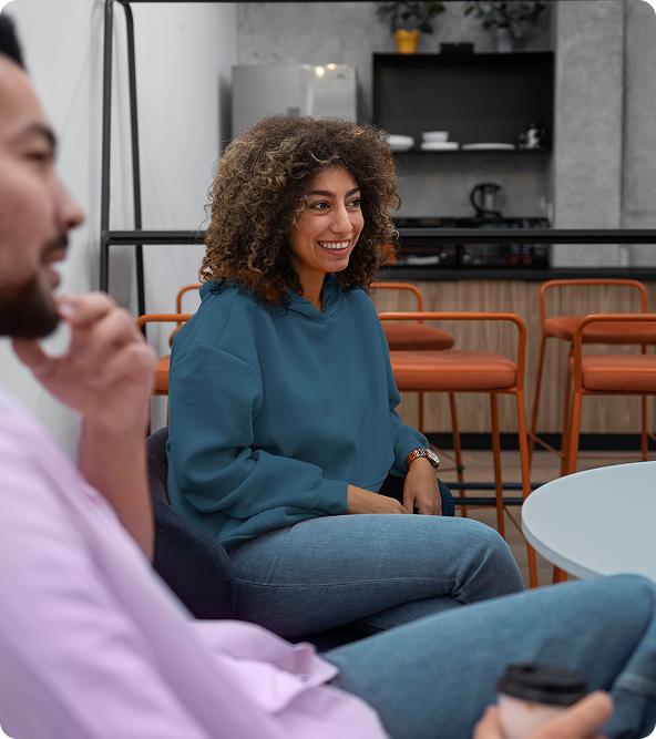 Woman in teal hoodie smiles, talking to someone off-screen in a modern cafe setting.