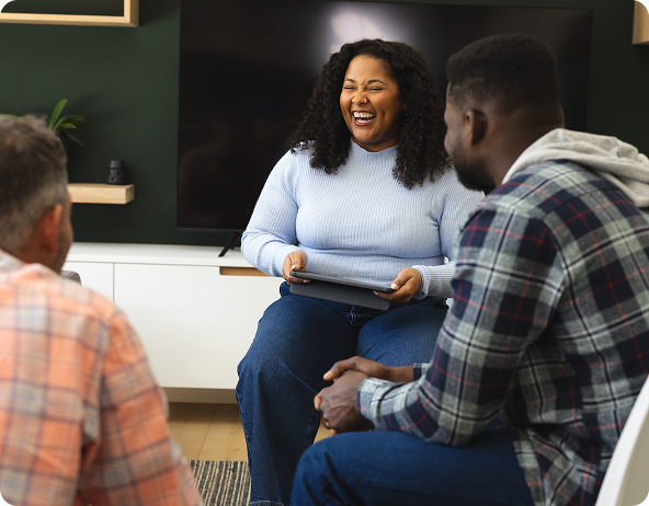 Three people in a counseling session. Woman laughs while holding a tablet. Two men listen.