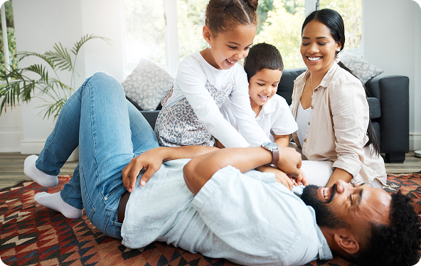 Family laughing and tickling each other on a rug in a living room.