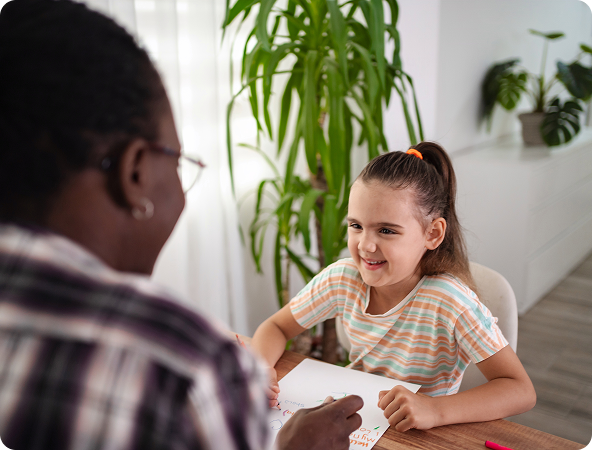 A person with glasses smiles at a girl seated at a table, both looking at paper; a plant is in the background.