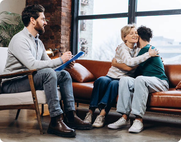 Therapist in session: Couple hugging on couch, therapist taking notes. Bright room with window.