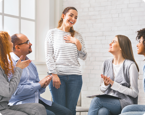 A woman smiles, center, in a group therapy setting, others clap.