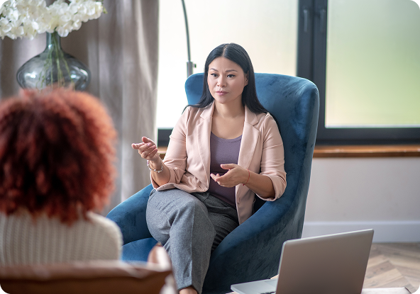 Woman in blazer gestures, talking to someone in a blue armchair; a laptop sits nearby.