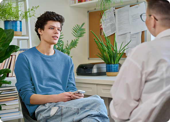 Teen in blue sweater talking to someone in a white coat indoors, plants in the background.