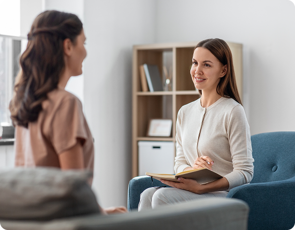 Two women in therapy session, one seated with a notebook, the other facing her, smiling in a room.